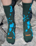 A person sitting on gray rocks wearing dark green socks with bright blue abstract shapes and orange TRAKK branding near the toes. 2